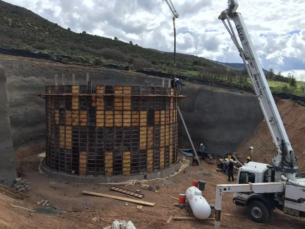 Crane truck pouring concrete into circular formwork for a property water cistern, mountain landscape backdrop.