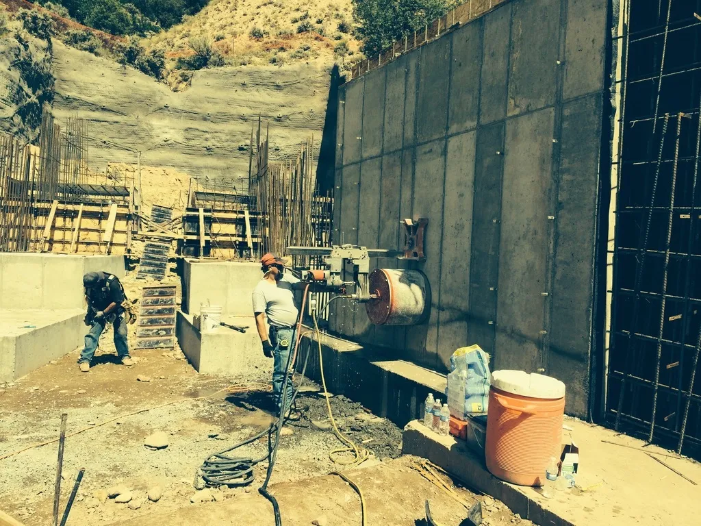 Workers at the base of a tall cast-in-place concrete retaining wall against a hillside cut, exposed rebar above.