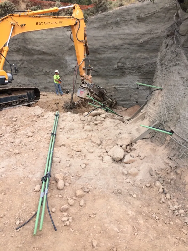 Drilling rig from B&Y Drilling working on a cut hillside, installing rock anchors or micropiles. Steel guide rods staged in the foreground.