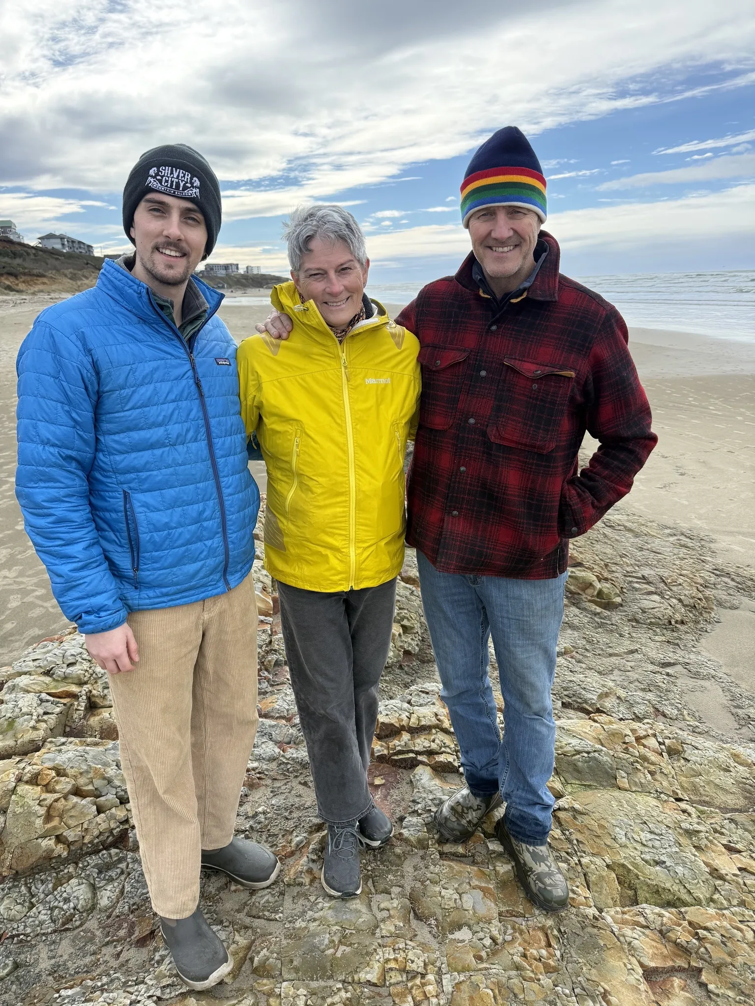 Marty Kribs (right) with his wife Blaire (center, yellow jacket) and son Wesley (left, blue jacket) on the beach in Newport, Oregon. All three in fleeces and beanies, Pacific Ocean behind them.