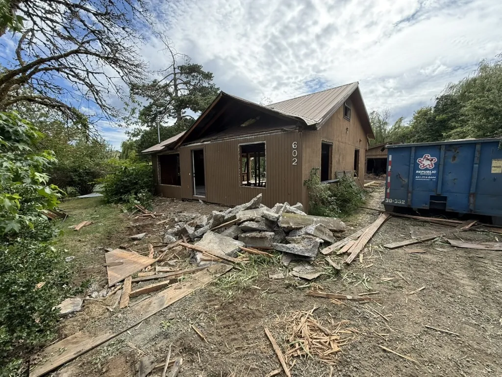 The Philomath property mid-demolition. Brown weathered siding, broken concrete and wood debris in a pile in the foreground, Republic Services dumpster, structural openings exposed.