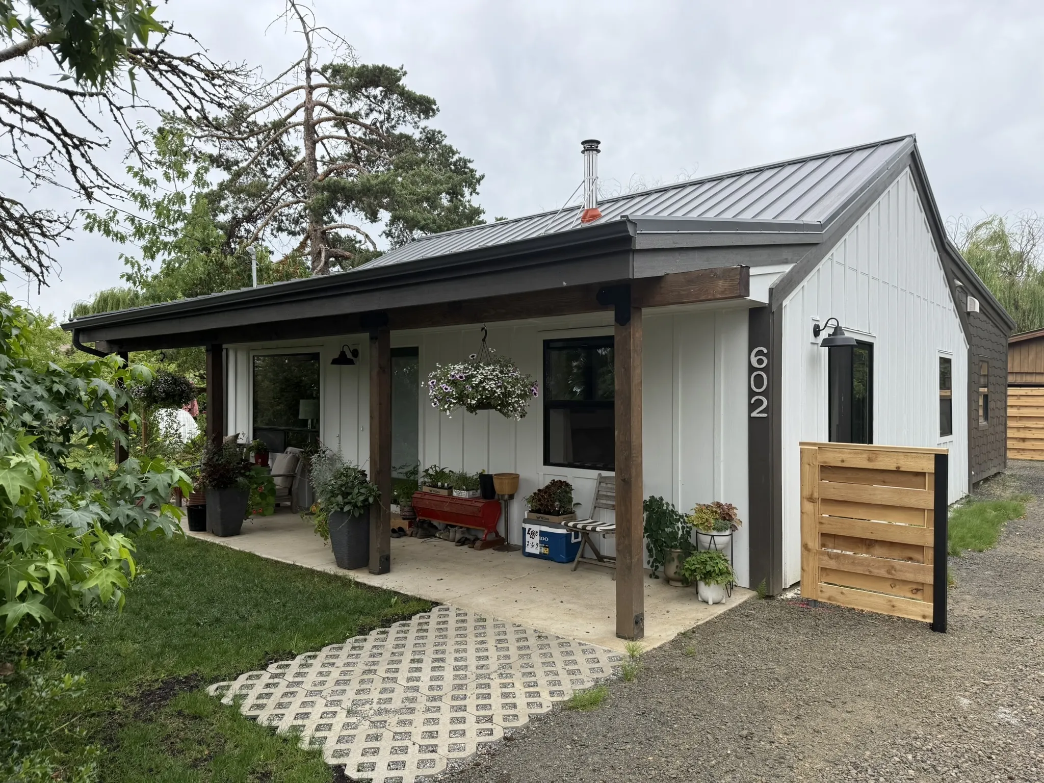 Finished Philomath, Oregon home. White board-and-batten siding, dark standing-seam metal roof, cedar porch posts, permeable paver walkway, garden plants in pots on the porch. Address number 602 visible.
