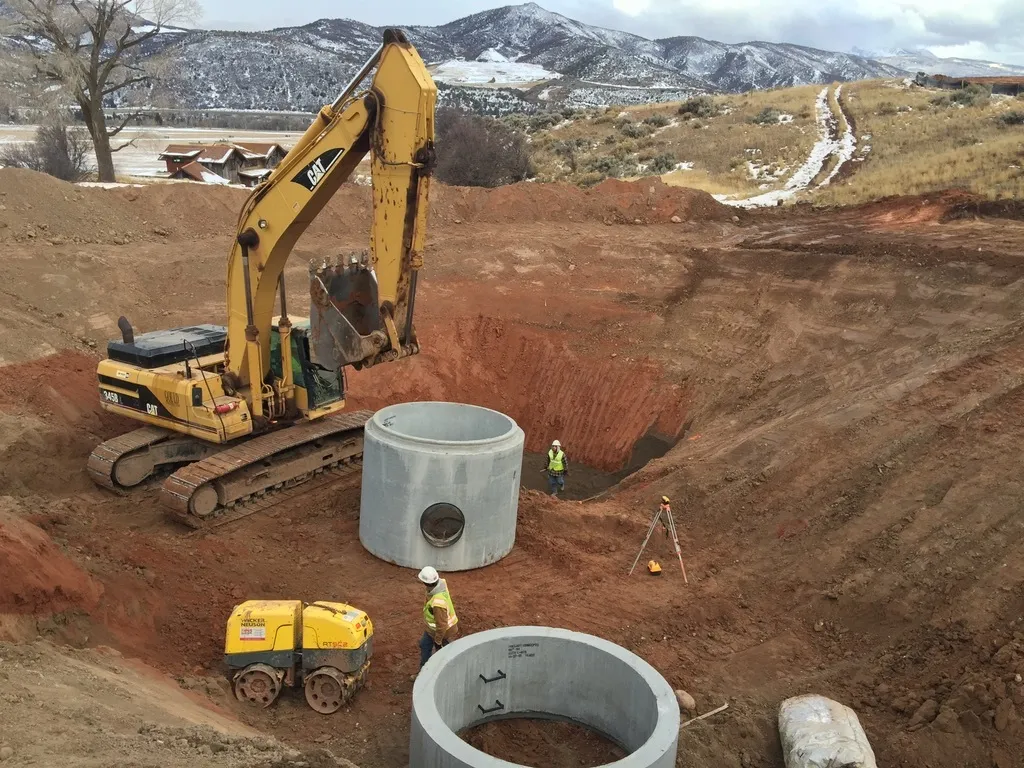 Excavator placing precast concrete cisterns on a snowy ranch property with snow-capped mountains and distant houses.